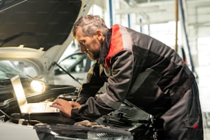 Middle aged male worker of car maintenance service bending over open engine compartment while looking at laptop display in workshop