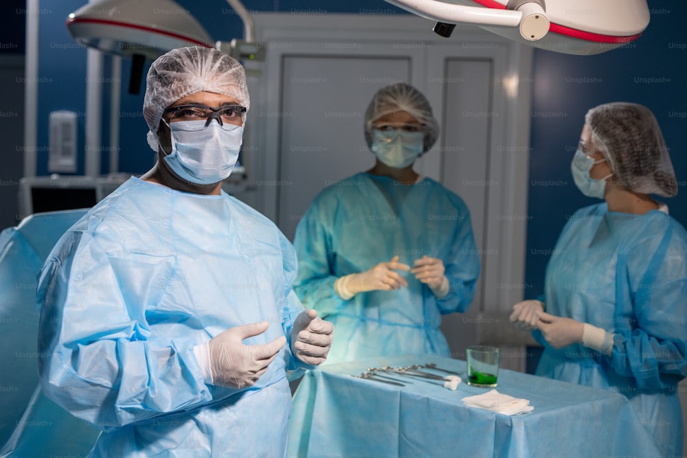 Contemporary mixed-race surgeon in protective mask, gloves and eyeglasses looking at you against his two female assistants
