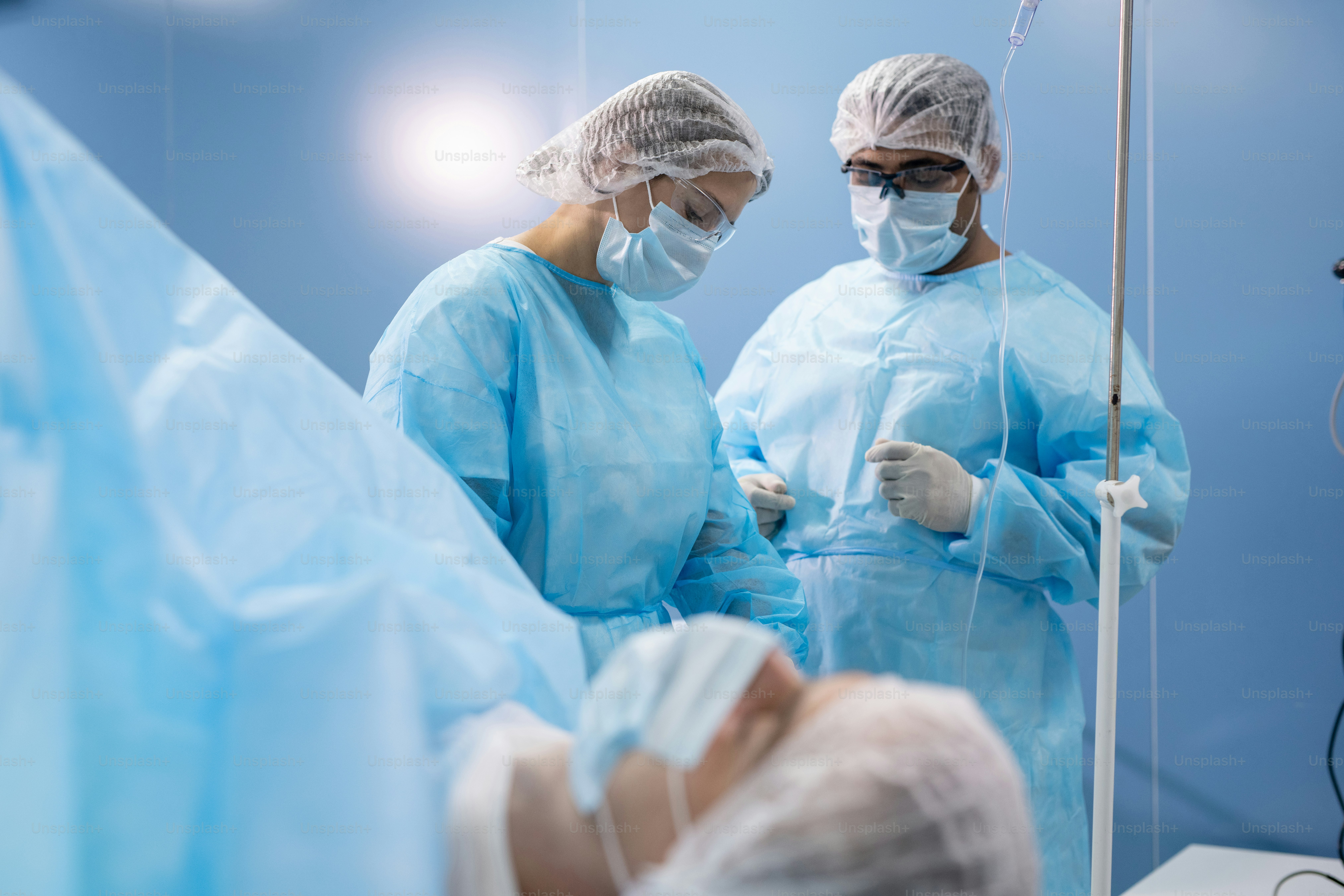 Young female assistant in protective workwear preparing medical instruments before operation between male surgeon and patient