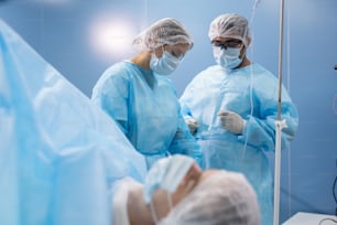 Joven asistente femenina en ropa de trabajo protectora que prepara instrumentos médicos antes de la operación entre el cirujano masculino y el paciente