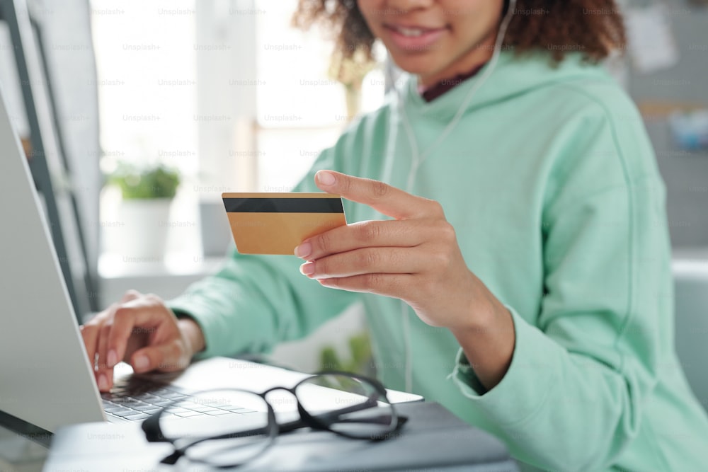 Mãos de jovem mulher mestiça em casualwear sentado na frente do laptop e indo pagar com cartão de crédito para compra na loja on-line