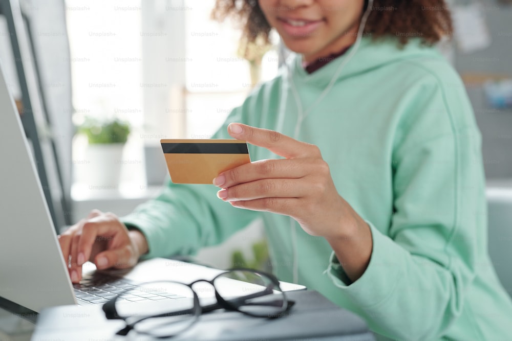 Mains d’une jeune femme métisse en tenue décontractée assise devant un ordinateur portable et allant payer par carte de crédit pour l’achat dans la boutique en ligne