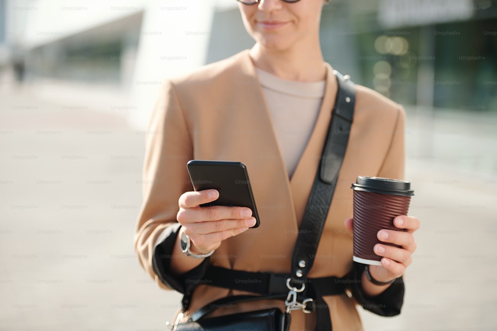 Hands of young businesswoman with glass of coffee scrolling through contacts in smartphone outdoors while going to phone someone