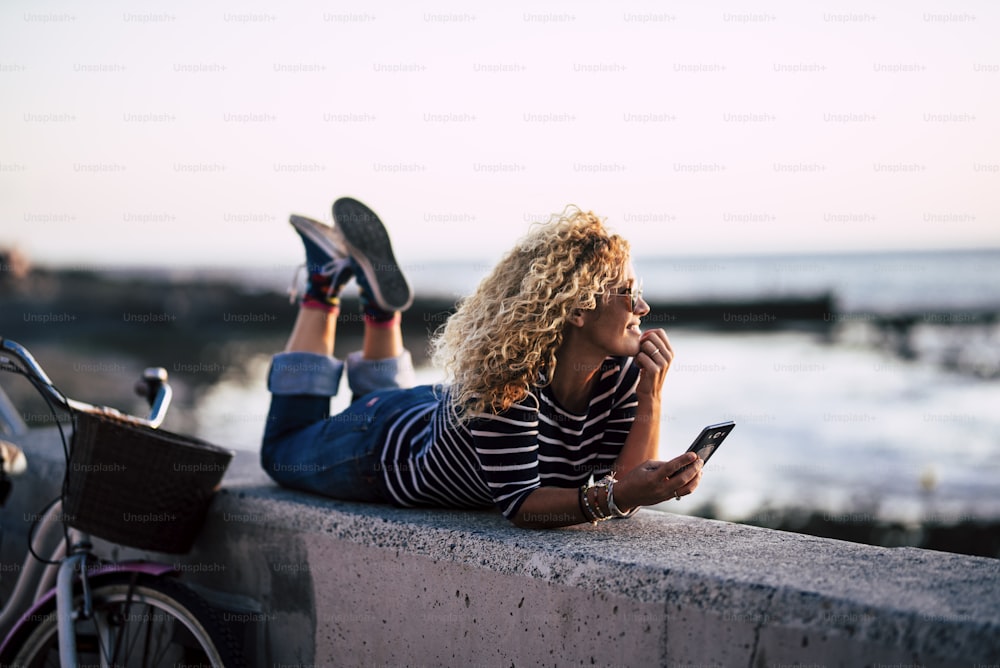 Happy beautiful blonde long curly hair woman enjoy the sunset lay down on the ground with blue ocean and sky in background - outdoor leisure activity for mature pretty people