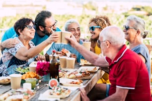 Group of different ages people celebrate and eat together in friendship outdoor at home - happy young, adult and senior have fun clinking and enjoying food on a wooden table - celebration and cheerful family friends