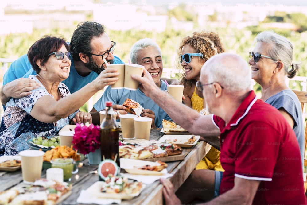 Groupe d’âges différents, les gens célèbrent et mangent ensemble dans l’amitié en plein air à la maison - jeunes, adultes et personnes âgées heureux s’amusent à trinquer et à déguster de la nourriture sur une table en bois - célébration et amis de la famille joyeux