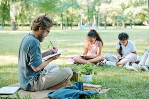 Profesor barbudo concentrado con gafas sentado con las piernas cruzadas y tomando notas en el portapapeles mientras los estudiantes hacen tareas en el parque