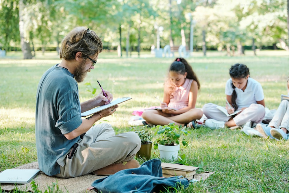 Professor barbudo concentrado em óculos sentado com as pernas cruzadas e fazendo anotações na prancheta enquanto os alunos fazem tarefas no parque