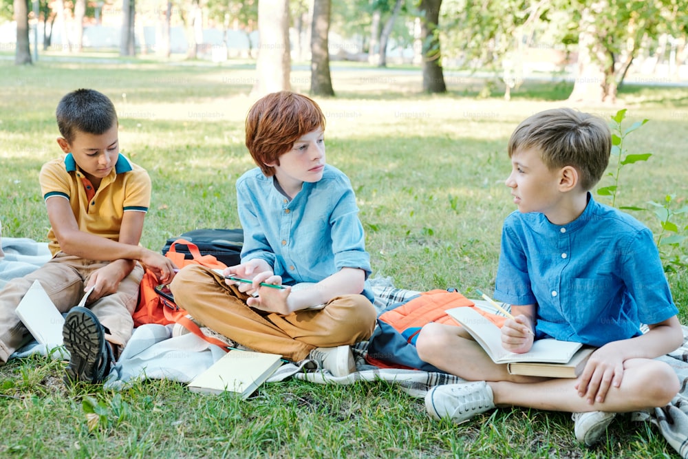 Grupo de niños pensativos sentados con libros en la hierba y discutiendo el proyecto escolar al aire libre