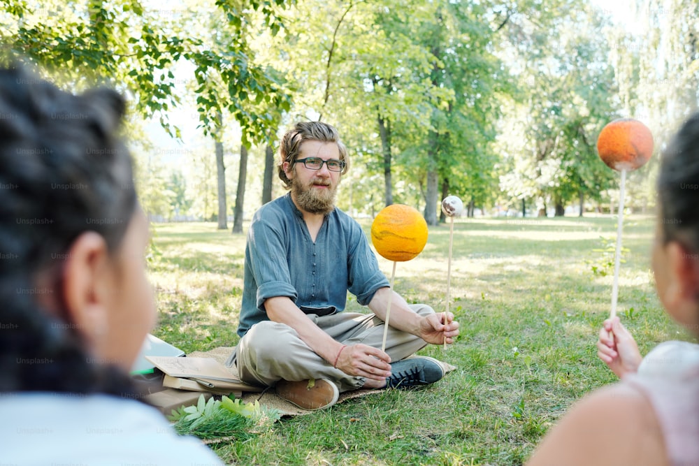 Smart bearded teacher sitting on ground and holding planet models while asking kids to describe it at outdoor lesson