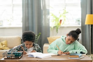 Cute diligent girl and boy in casualwear making notes in copybooks while sitting by table in living-room against armchairs and large windows