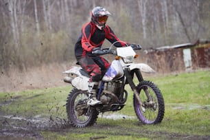 Motorcross rider racing in mud track against dry trees and green lawn