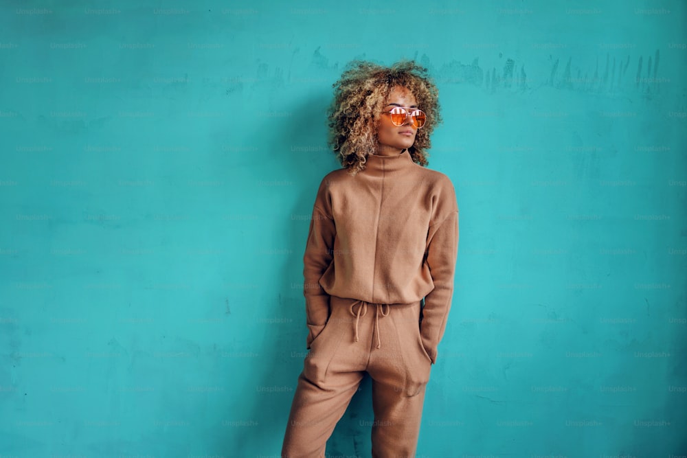 Young fashionable woman with curly hair holding hands in pockets and standing in front of turquoise wall.