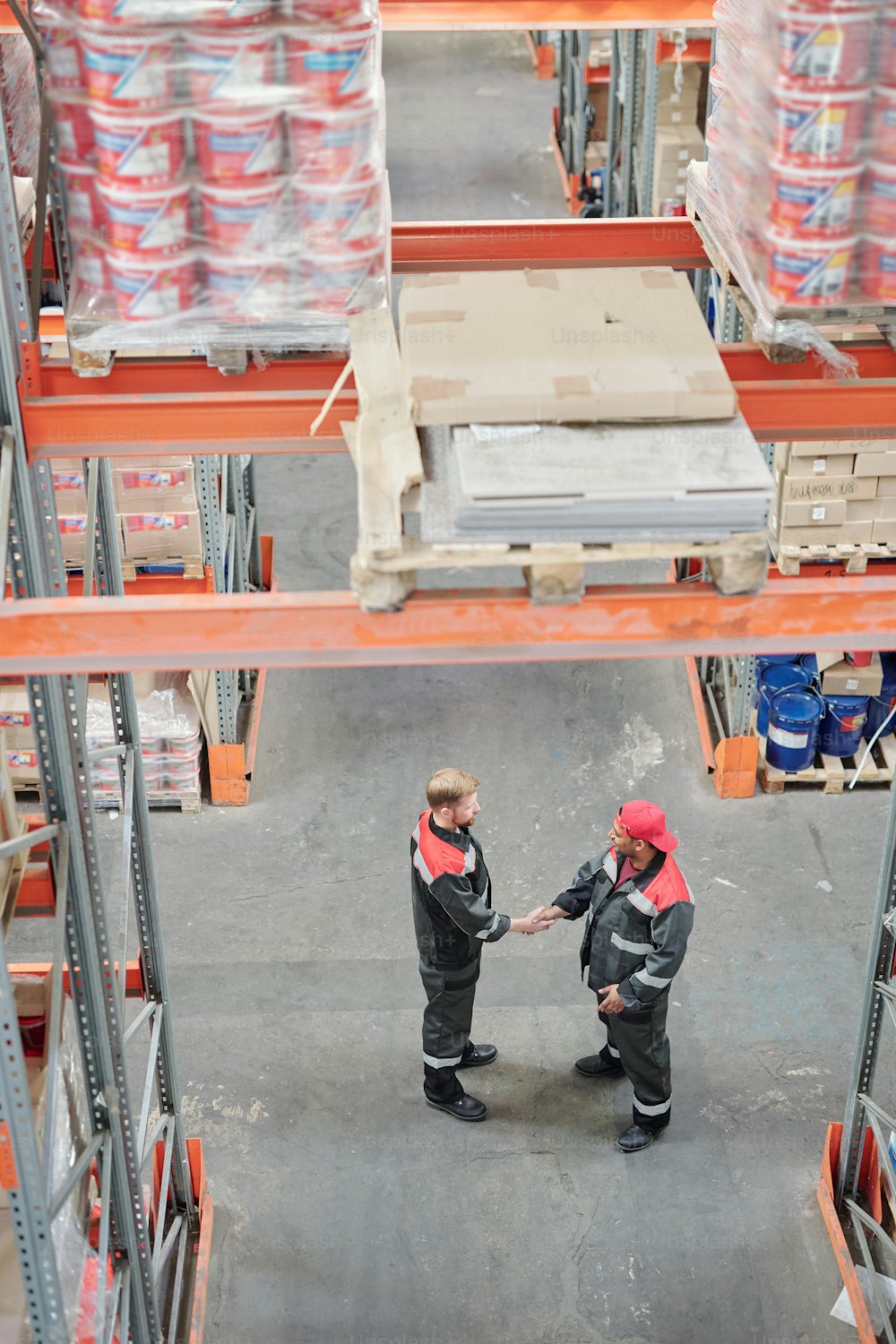 Two young successful intercultural male managers of warehouse hall shaking hands while greeting each other on aisle between racks