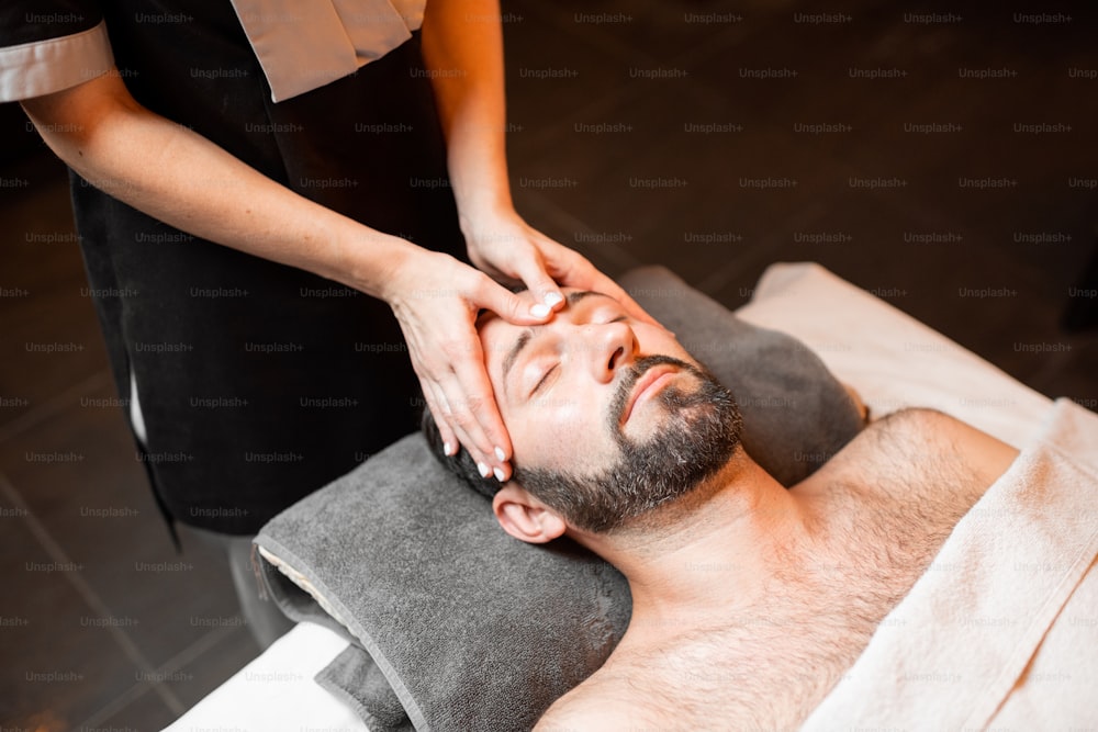 Hombre barbudo recibiendo un masaje facial, relajándose en el salón del balneario, primer plano