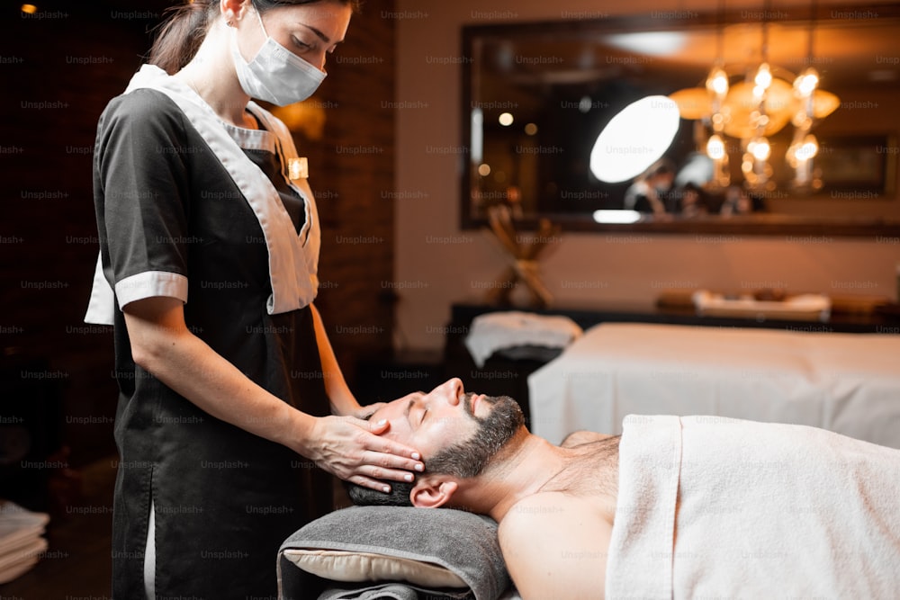 Masajista profesional con mascarilla médica haciendo masaje facial a un cliente masculino en el salón Spa. Negocios durante el concepto de epidemia