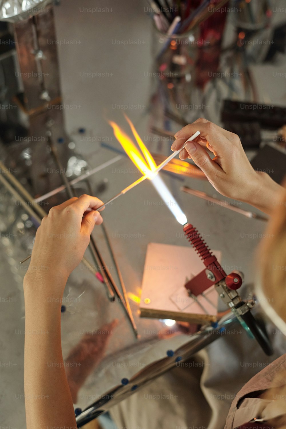 Mani di giovane artigiana che bruciano il pezzo lungo a forma di bastone di vetro mentre lo tengono sul fuoco uscendo dal bruciatore durante la lavorazione a lume