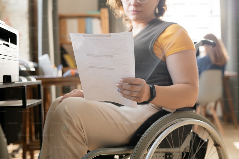 Hand of young contemporary businesswoman reading paper while sitting in wheelchair and going to make copy on xerox machine in office