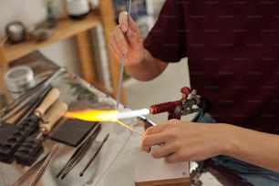 Hände einer jungen zeitgenössischen Kunsthandwerkerin, die am Arbeitsplatz sitzt und das Werkstück mit Feuer verbrennt, während sie es in der Werkstatt über den Brenner hält