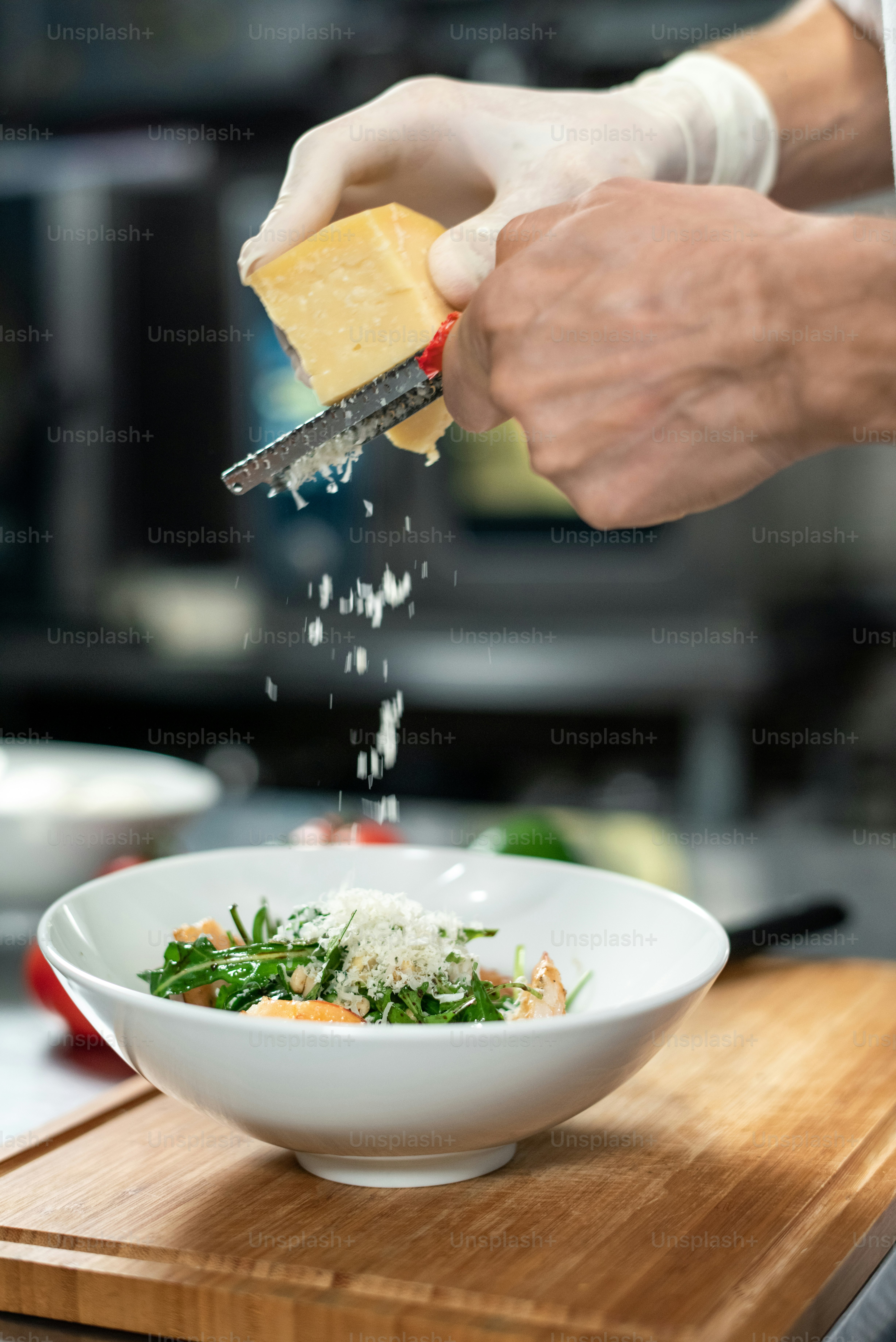 Hands of young chef grating fresh cheese into ceramic bowl containing salad consisting of ruccola, boiled shrimps and other ingredients