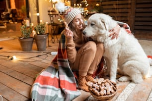 Retrato de una mujer con su lindo perro celebrando unas vacaciones de Año Nuevo, sentados juntos y comiendo panes de jengibre en la terraza bellamente decorada en casa