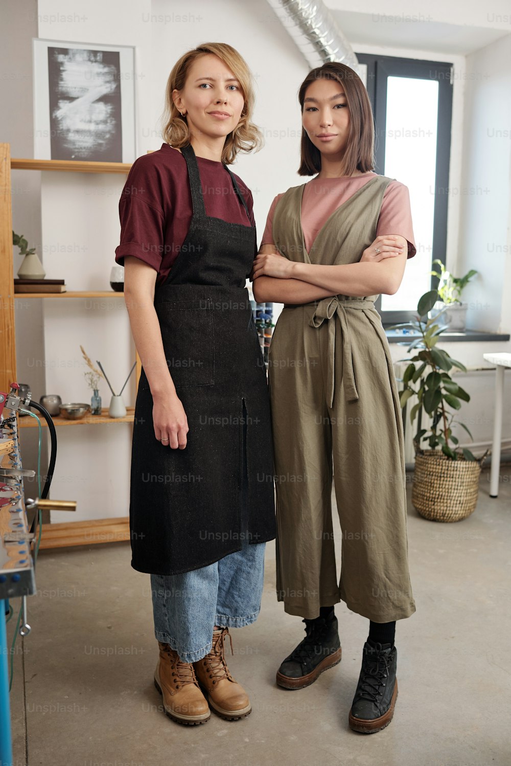 Two young intercultural female artisans in workwear standing next to one another in front of camera in large workshop and looking at you