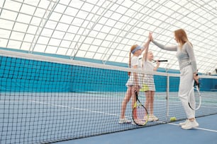 Des préadolescentes heureuses en tenue de sport donnent des high five à leur jeune entraîneur de tennis sur le filet avant ou après l’entraînement dans un stade moderne