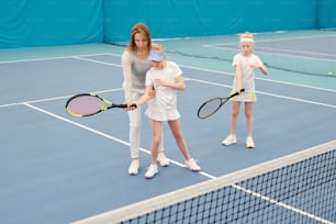 운동복을 입은 젊은 테니스 트레이너가 경기장에서 개별 훈련 중에 그녀와 상담하는 동안 두 명의 귀여운 소녀 중 한 명이 들고 있는 라켓을 들고 있습니다.