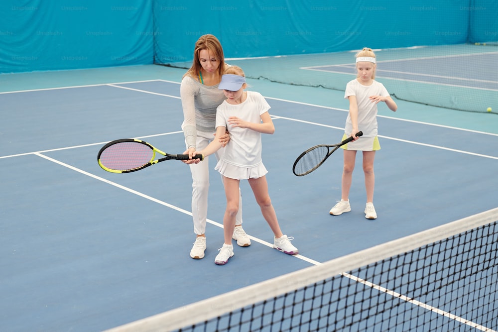 Giovane istruttore di tennis in abbigliamento sportivo che tiene la racchetta tenuta da una delle due ragazze carine mentre la consulta durante l'allenamento individuale sullo stadio