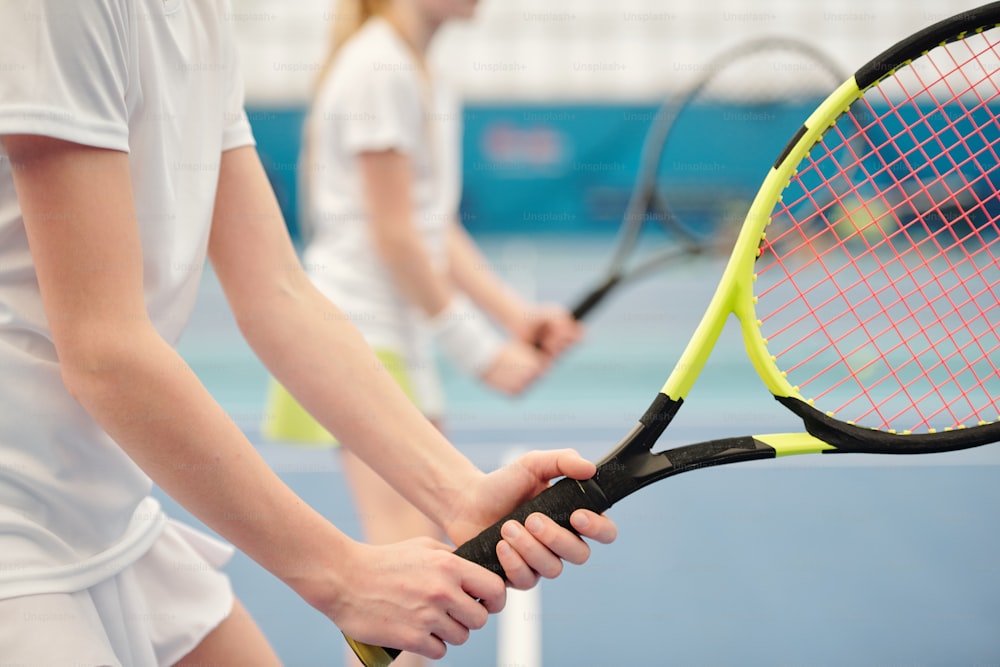 Manos de una adolescente en ropa deportiva blanca de pie en el estadio contra el campo y sosteniendo una raqueta de tenis lista para empujar la pelota durante el juego