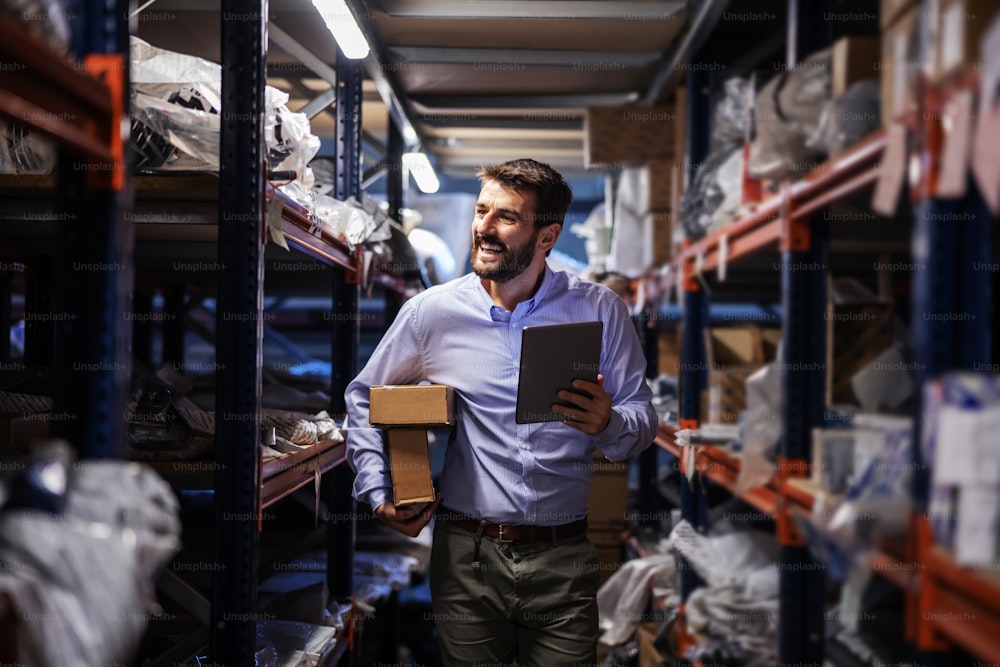 Smiling bearded businessman walking trough storage of shipping firm, holding boxes under armpit and using tablet to check on goods.