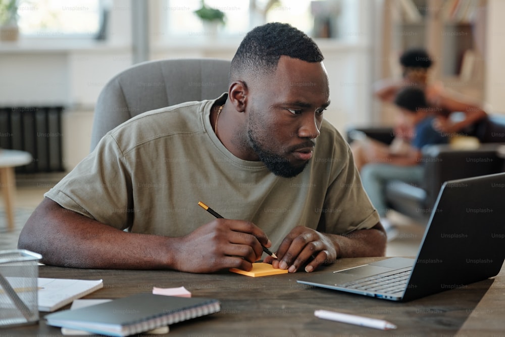 Young African man with pencil over notepaper sitting by table in front of laptop and looking through online data against his wife and little son