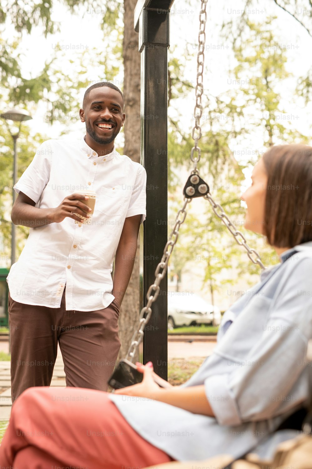 Happy African guy with drink standing in front of young woman relaxing on swing while talking to her during chill on summer day in park