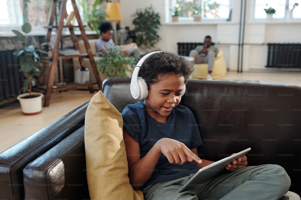 Adorable little boy with headphones sitting on black leather couch by pillow and pointing at screen of digital tablet against his parents