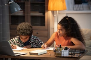 Dos lindos hermanos de etnia africana sentados junto a la mesa en casa y tomando notas en cuadernos frente a la computadora portátil mientras ven la lección en línea