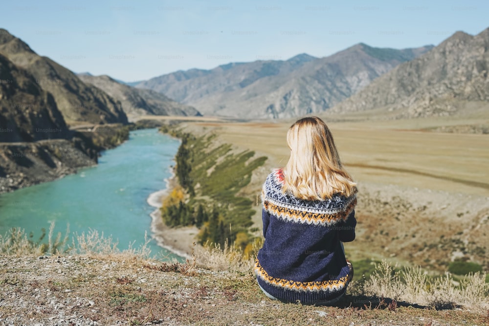 청록색 카툰 강, 알타이 산맥의 배경에 앉아 북유럽 스웨터를 입은 젊은 금발 여자