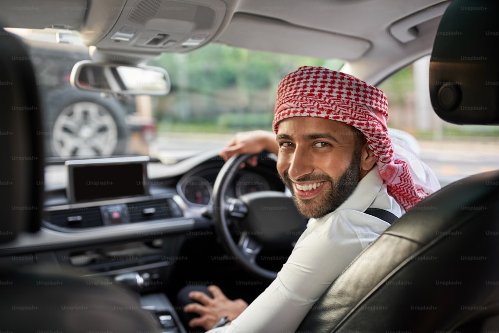 Bel uomo arabo che guarda indietro sorridendo ai passeggeri sul sedile posteriore del suo taxi ehailing. Uomo arabo che indossa la kefiah tradizionale del velo