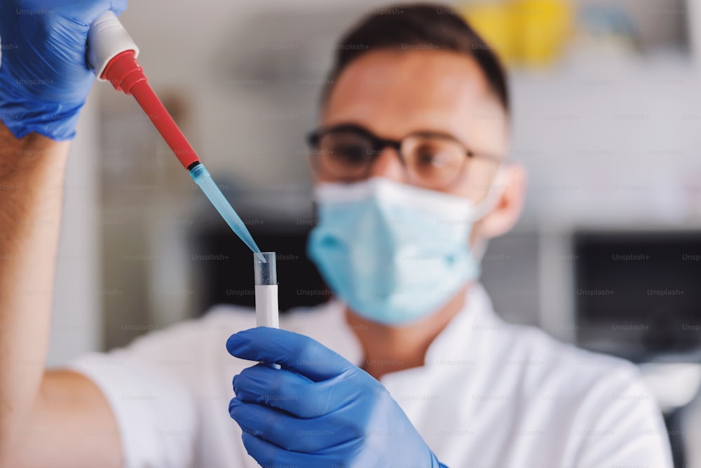 Atraente assistente de laboratório masculino com luvas de borracha e máscara facial segurando tubo de ensaio com sangue e fazendo pesquisas para a cura do corona vírus.