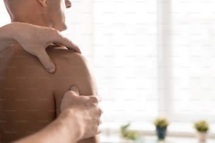 Mains d’un masseur professionnel ou d’un physiothérapeute massant l’épaule d’un patient masculin mature torse nu assis devant lui dans les cliniques