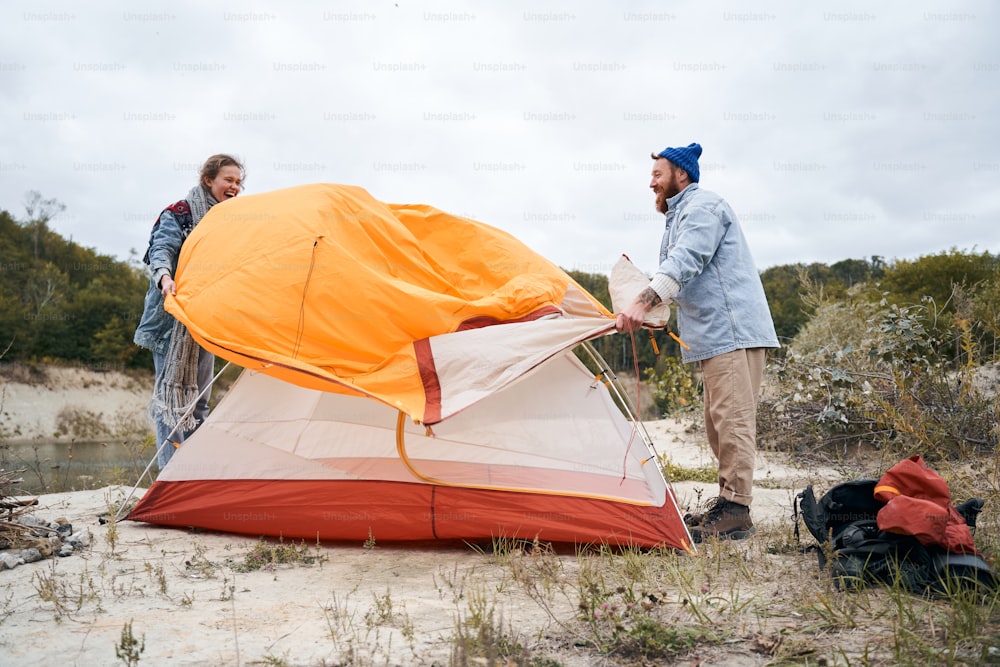 Divirta-se juntos! Menina engraçada rindo em voz alta enquanto ajudava seu amado homem a cobrir a tenda com uma camada protetora