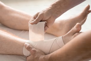 Mains d’un clinicien contemporain enveloppant le pied et la cheville d’un patient avec un bandage flexible pendant une procédure médicale sur un divan à l’hôpital