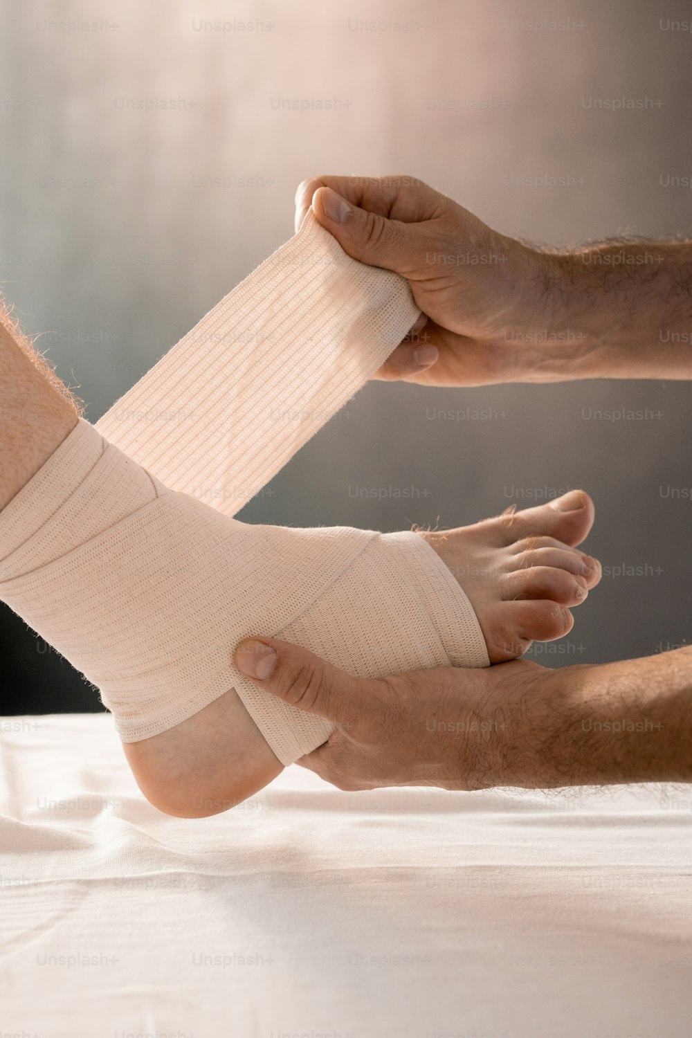 Hands of clinician holding foot of male patient with sick leg while wrapping it with flexible bandage during medical procedure in clinics