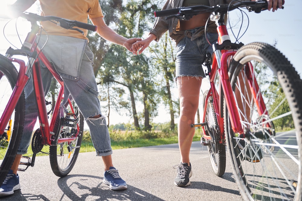 Cropped view of active young couple with bikes standing outdoors on a road in nature and holding hands before the start