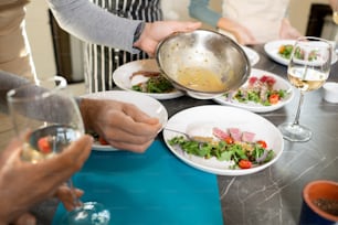Hands of mature man putting sauce from metallic bowl on plate with salad consisting of fresh cherry tomatoes, ruccola and chopped onions