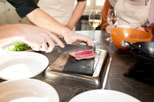 Hands of young male cooking coach chopping piece of smoked or grilled beef on special board by table with two trainees standing near by