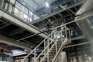 Bald mature foreman with digital tablet working with technical data inside contemporary large polymer processing factory in front of staircase