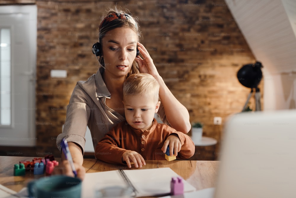 Mid adult working mother taking notes and communicating over headset while small son is sitting on her lap at home.
