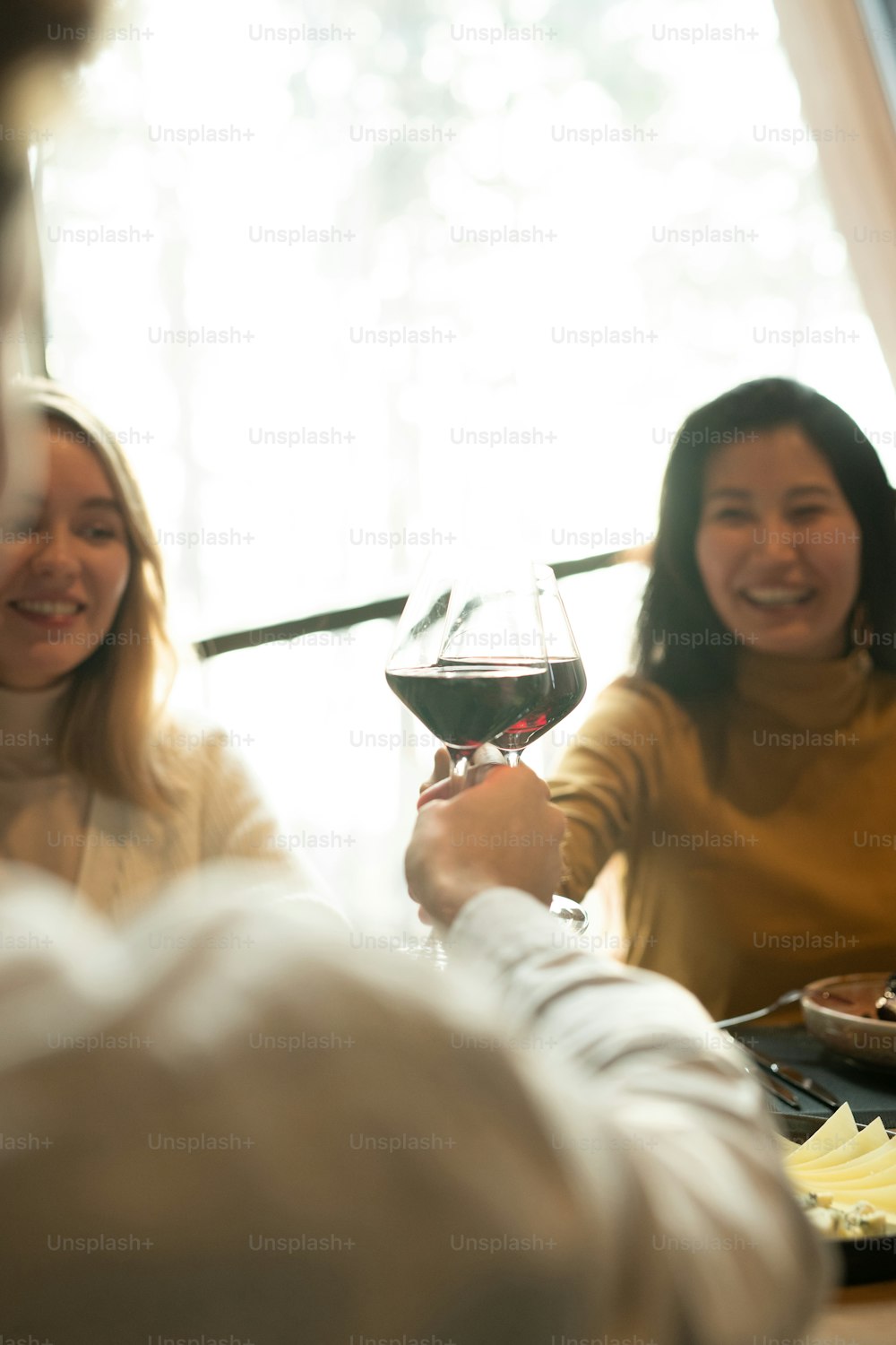 Young cheerful Asian woman clinking with glass of red wine with her boyfriend among friends over served table by dinner in restaurant