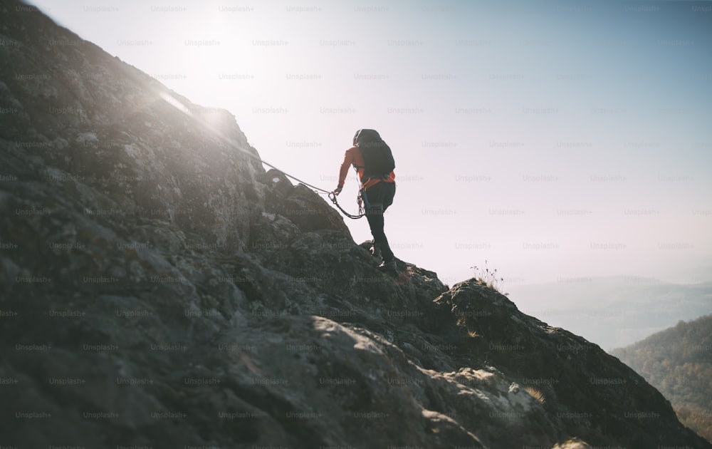Montañero con mochila que usa cuerda de escalada para escalar la cumbre de la montaña rocosa.