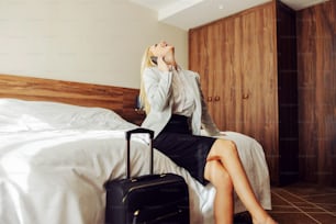 Laughing middle-aged woman sitting on the bed in a hotel room and having a phone conversation with someone. Next to her is a suitcase.